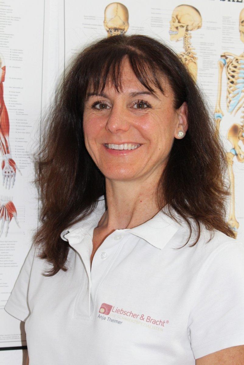 Anja Theimer, Physiotherapeutin und sktorale Heilpraktikerin aus Ladenburg steht vor Anatomie Plakaten. Sie hat lange wellige braune Haare mit Pony. Zudem trägt sie ein weißes Liebscher & Bracht Polo T-Shirt.